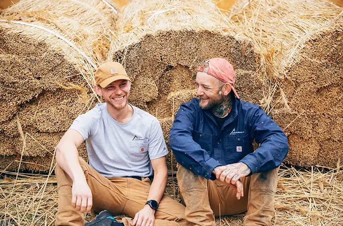 To smilende tækkemænd fra Tækkekollektivet i arbejdstøj sidder foran store halmballer og taler sammen i landlige omgivelser.
